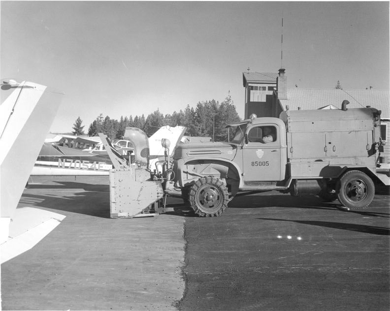 A utility truck with equipment is parked on an airfield near small airplanes.