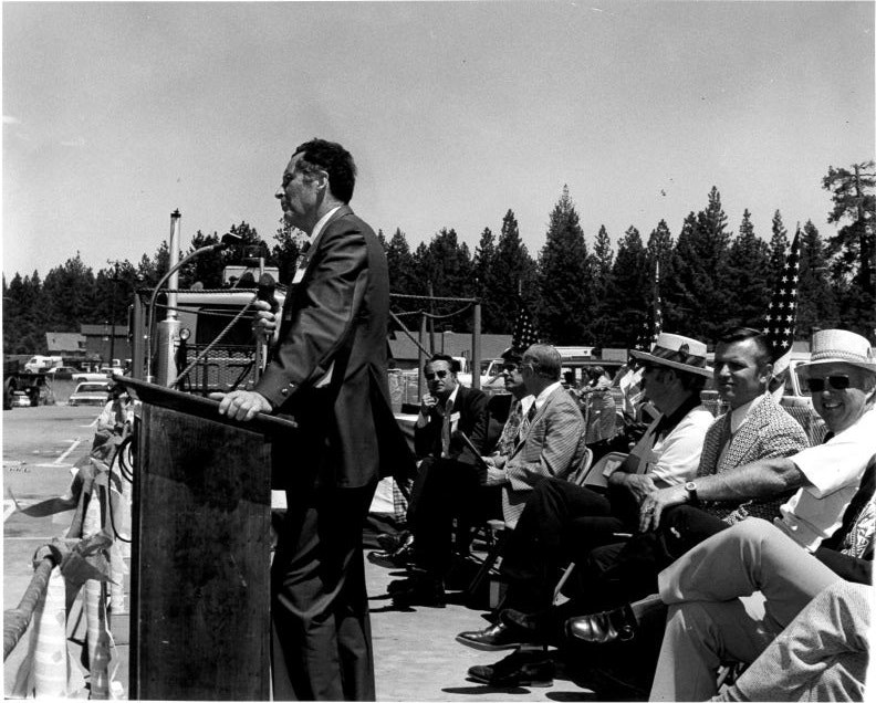 A man speaks at a podium outdoors, surrounded by seated people in suits and hats, with trees and cars in the background.