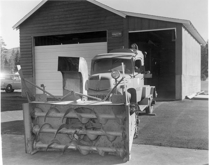 A snowplow truck parked outside a garage with a large snow auger attachment in front.