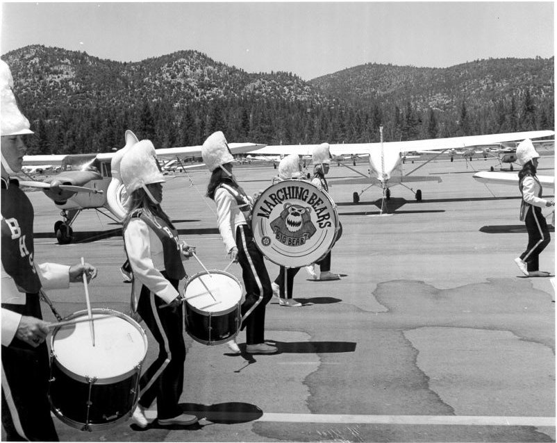 A marching band called "Marching Bears" performs on an airstrip with small airplanes in the background.