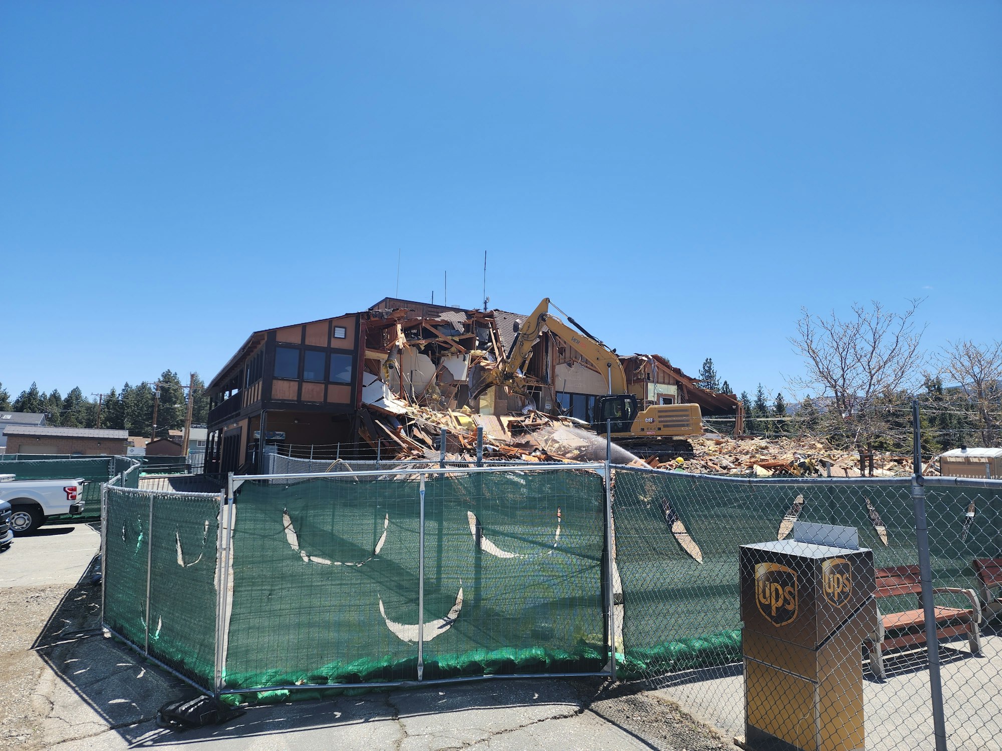 A partially demolished building with debris, an excavator, green fencing, and a UPS drop box nearby.