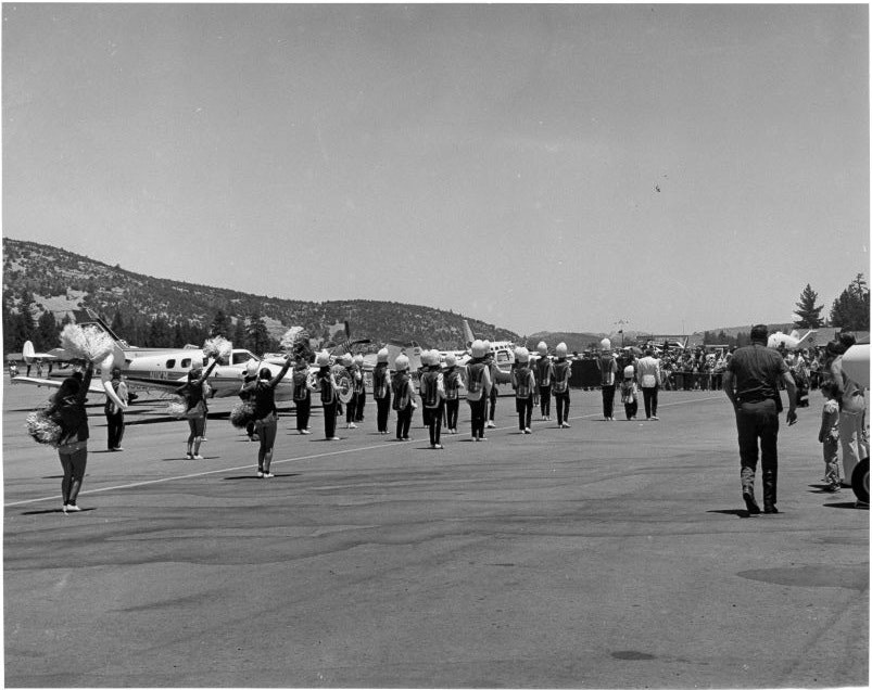 Marching band with pom-pom dancers performing on an airport tarmac with airplanes and spectators in the background.
