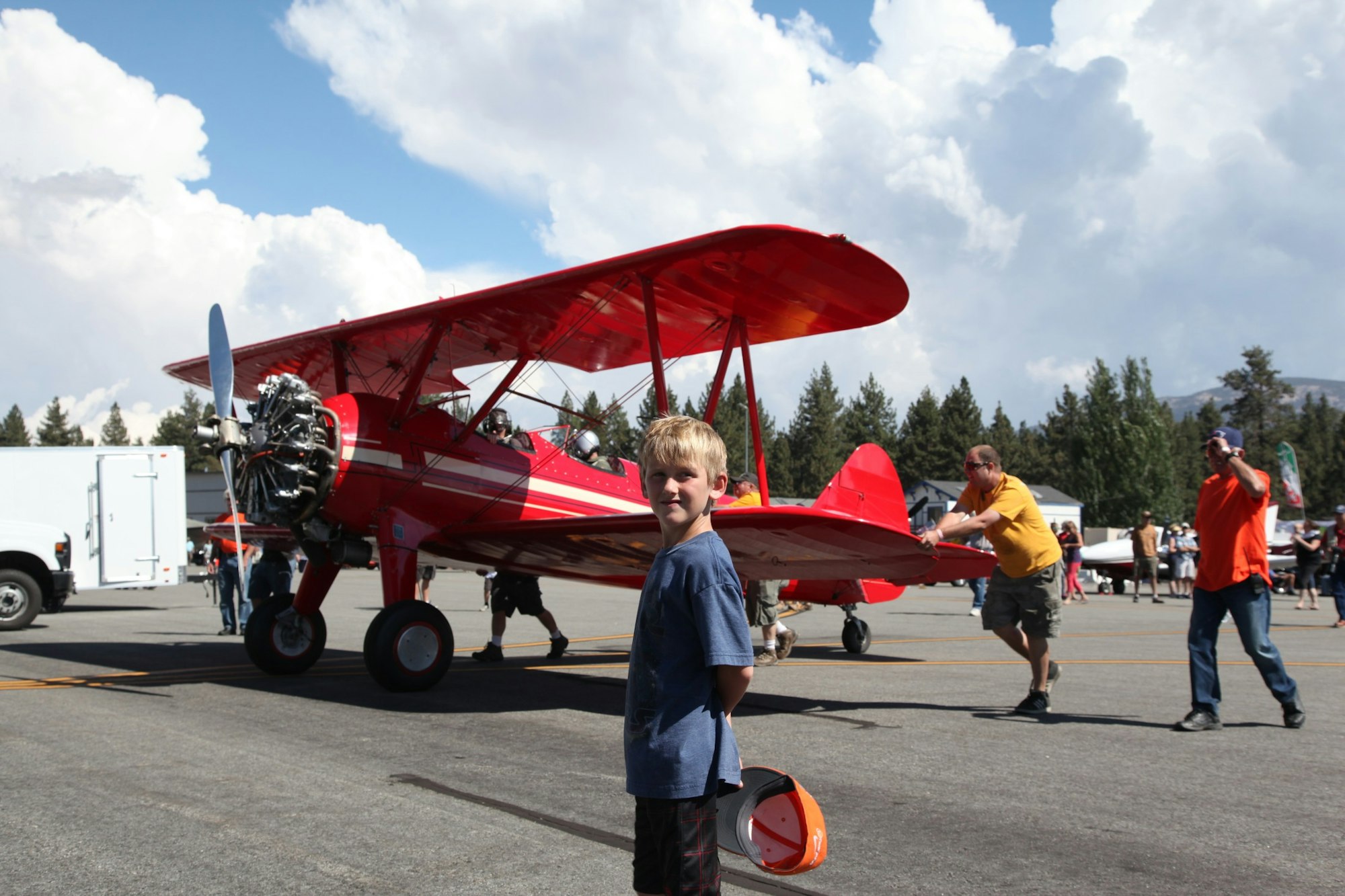 A boy stands near a red biplane at an airshow. People push the plane on the tarmac.