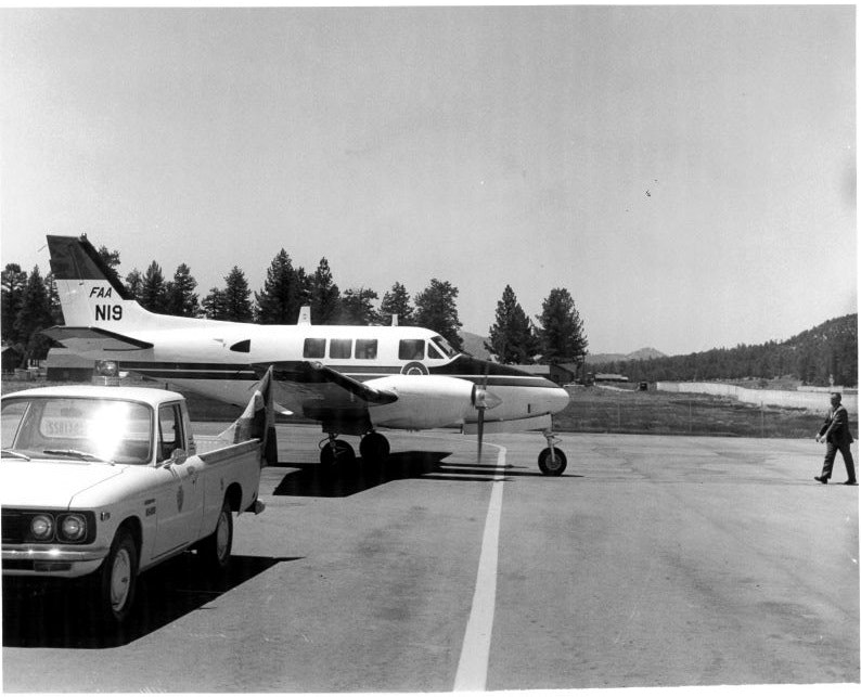 A small airplane marked "FAA N19" parked on an airfield with a truck nearby and a person walking towards it.
