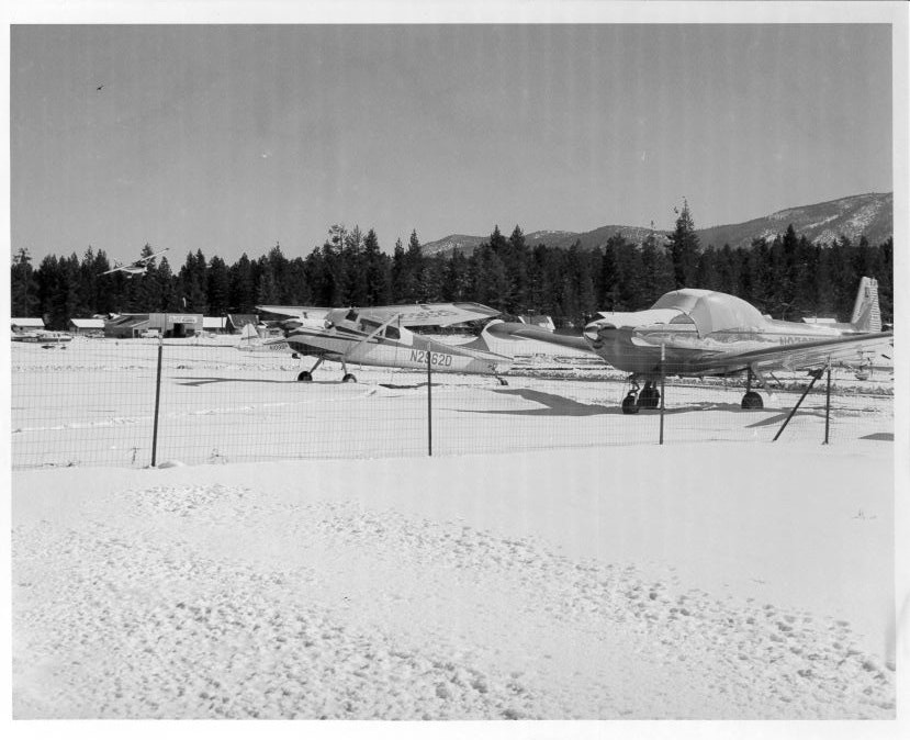 Two small airplanes parked on a snowy airfield, surrounded by trees and mountains in the background.