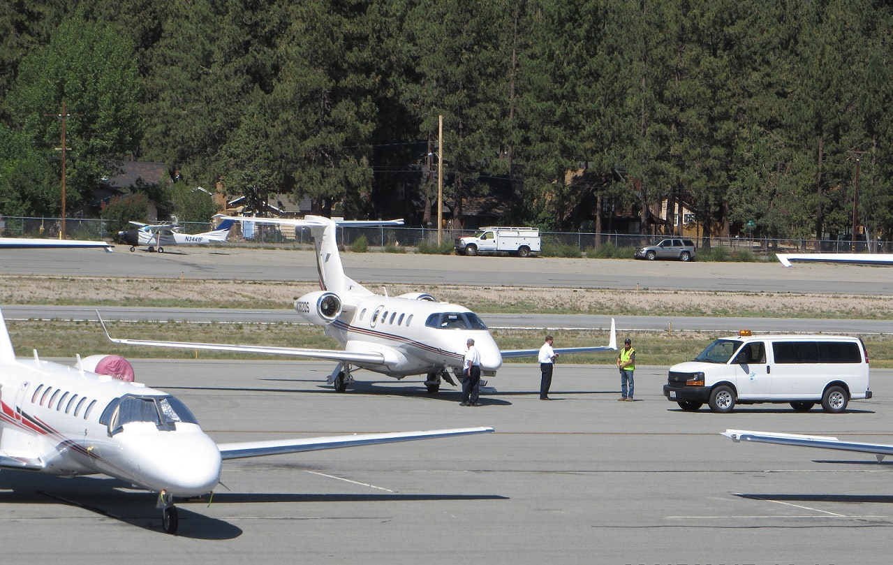 Several private jets on a runway, people standing nearby, with a white van and forested background.