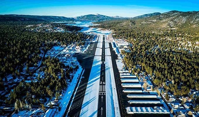 Aerial view of a snow-covered airport runway surrounded by forest and mountains.