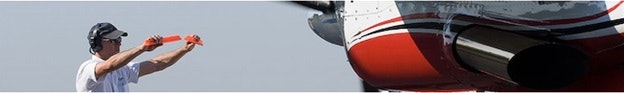A person with headphones and orange batons directs an aircraft on the ground.