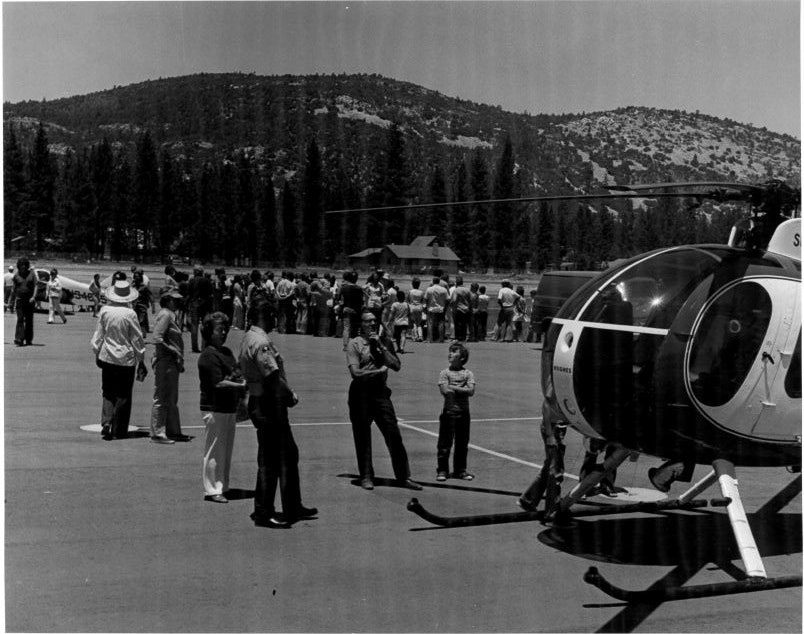 A crowd gathers near a helicopter on a tarmac with a forested hill in the background.