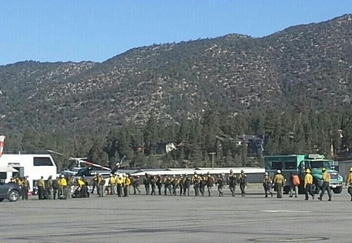 A group of people in yellow jackets near a helicopter and vehicles, set against a mountainous backdrop.