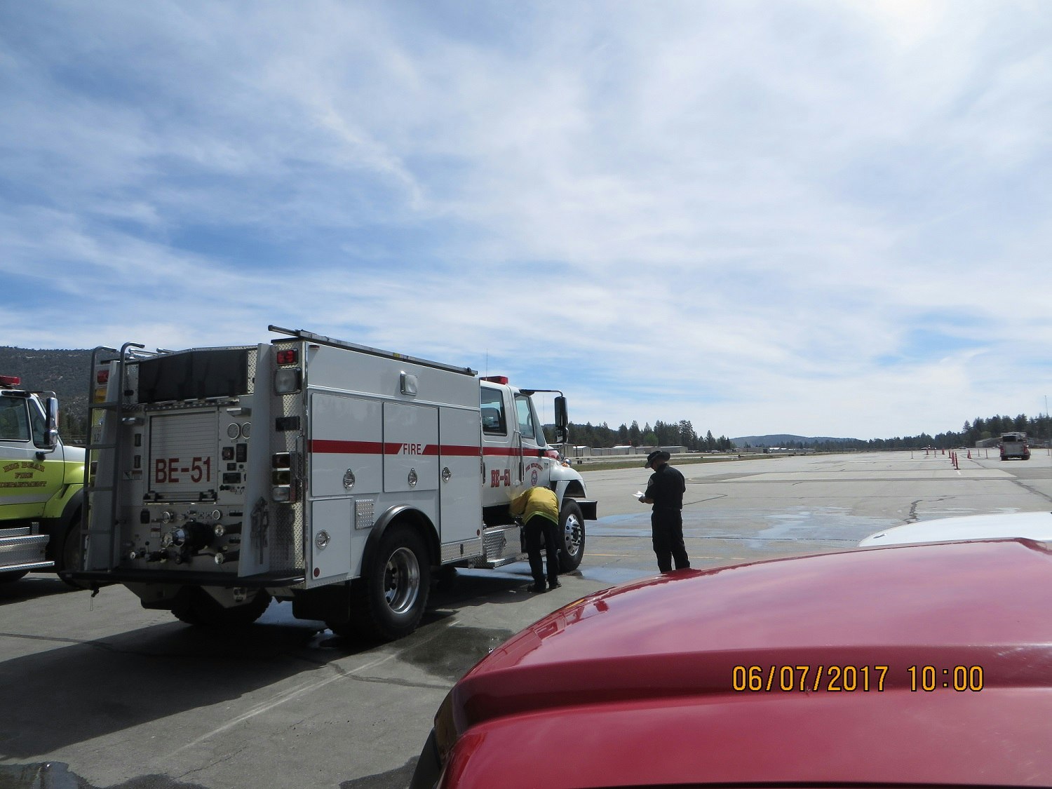 Two fire trucks on a paved area with two people near them under a partly cloudy sky.