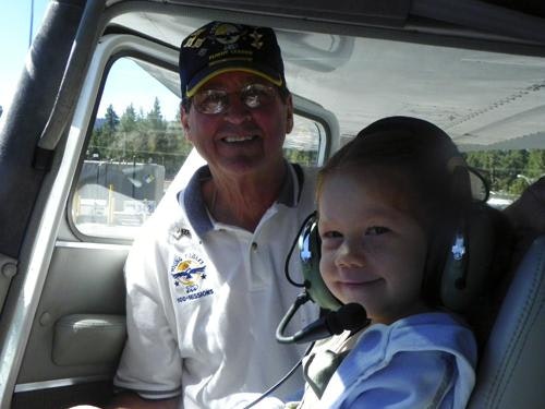 An older person and a child wearing headsets are sitting in the cockpit of a small aircraft, smiling at the camera.