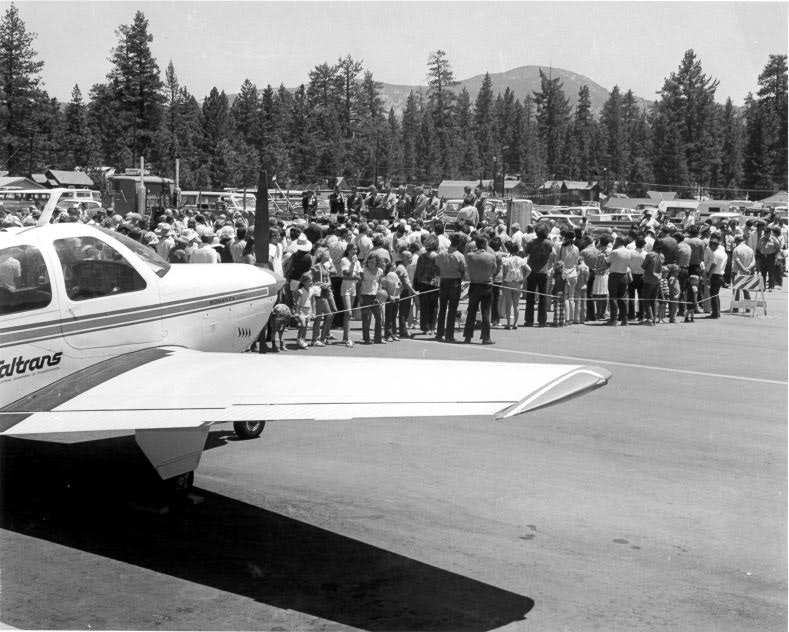 A crowd gathered near an airplane at an outdoor event surrounded by trees and mountains.