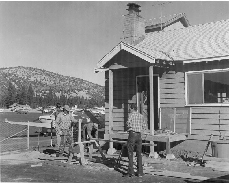Three people work on a house porch near an airport, with small airplanes and mountains in the background.