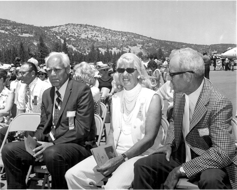 A group of people seated outdoors in formal attire, with a mountainous landscape in the background.