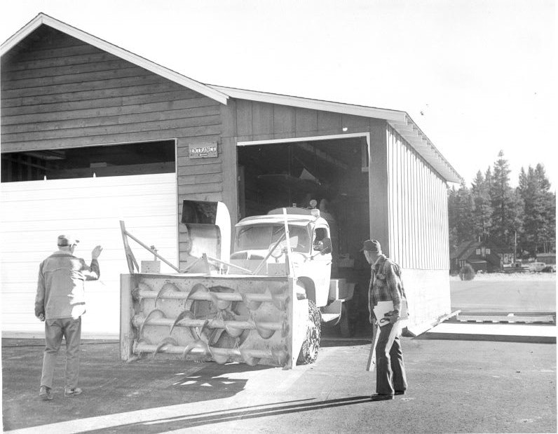A snowplow truck emerges from a garage as two people direct and observe.