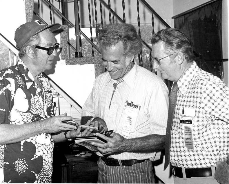 Three men in patterned shirts are engaged in conversation, looking at a notepad.