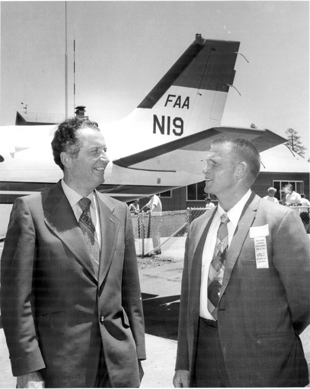 Two men in suits conversing in front of a small airplane with "FAA" and "N19" on the tail.