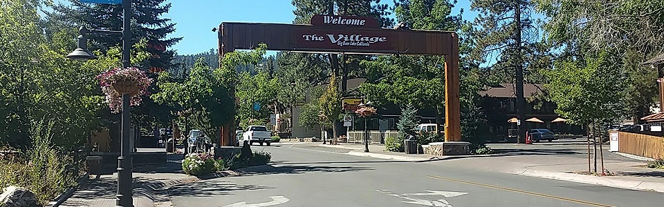 Street entrance with a "Welcome to The Village" sign, surrounded by trees and lampposts, in a sunny area.