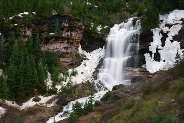 A cascading waterfall with remnants of snow, surrounded by evergreen trees and rocky terrain.