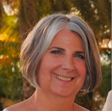 A smiling woman with short gray hair, outdoor background with trees.