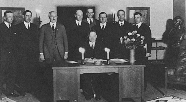 A historical black-and-white photo of a group of men in suits, one seated at a desk, possibly a formal gathering or signing event.