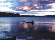 A person canoeing on a calm lake at dusk with a colorful sky.