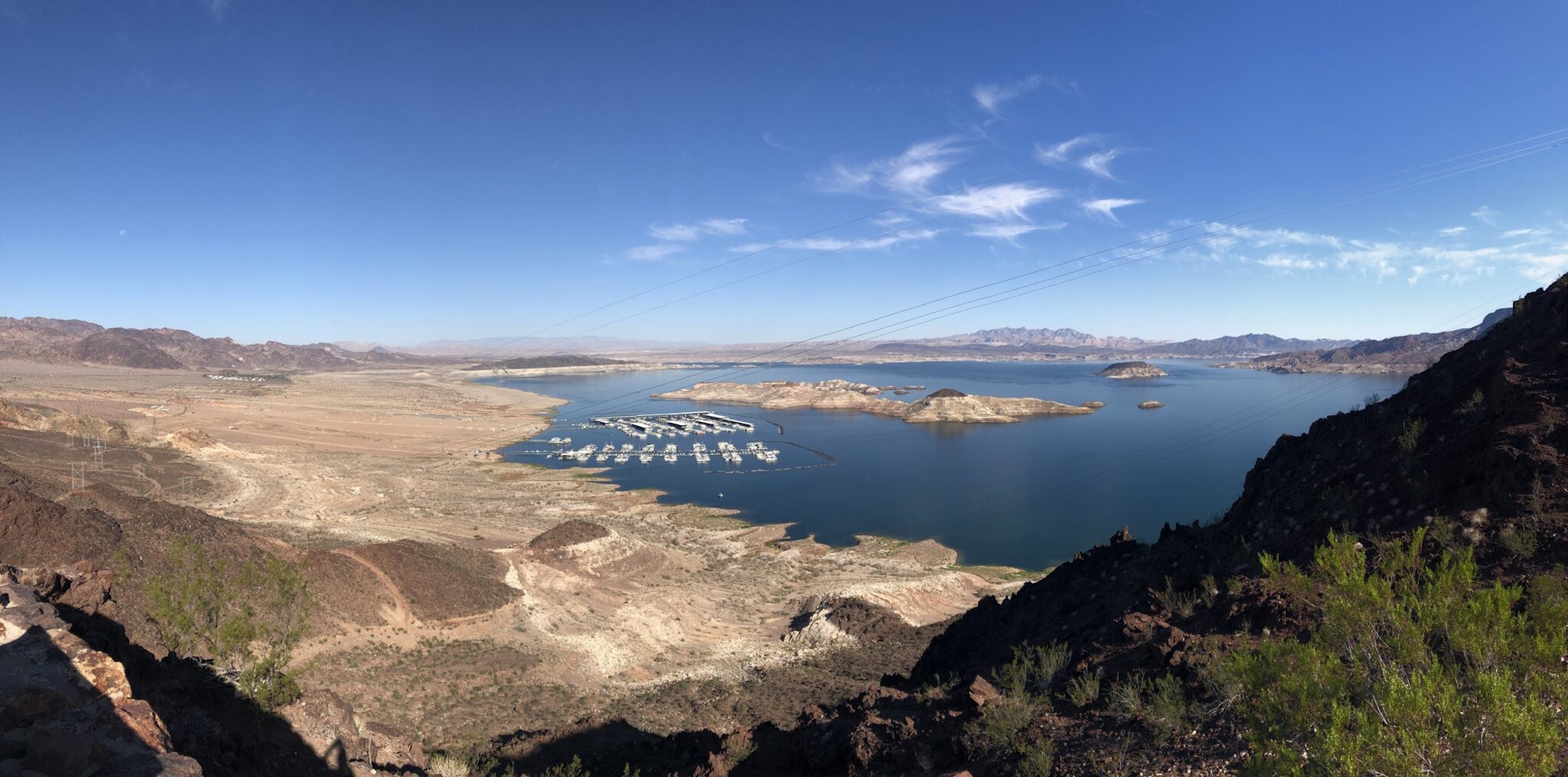 A panoramic view of a lake with boats, surrounding desert terrain, and a clear blue sky with wispy clouds.