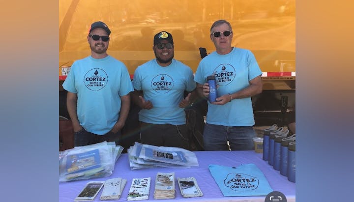 Three men in matching shirts stand behind a table with informational materials, possibly at an event.