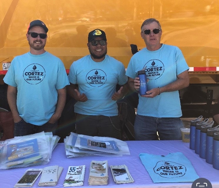 Three men in matching shirts stand behind a table with informational materials, possibly at an event.