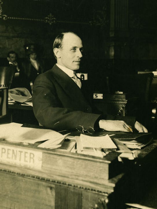 A vintage photo of a man at a desk with papers, a nameplate reading "CARPENTER," and eyeglasses in hand.