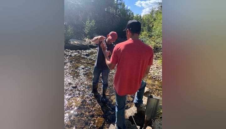 Two people by a stream, one holding a fish they've caught. Trees and sunlight visible.