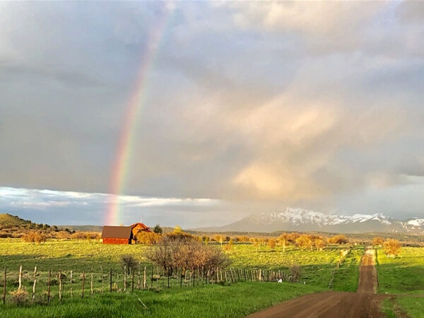 A rainbow over a rural landscape with a red barn, a dirt road, and mountains in the background.