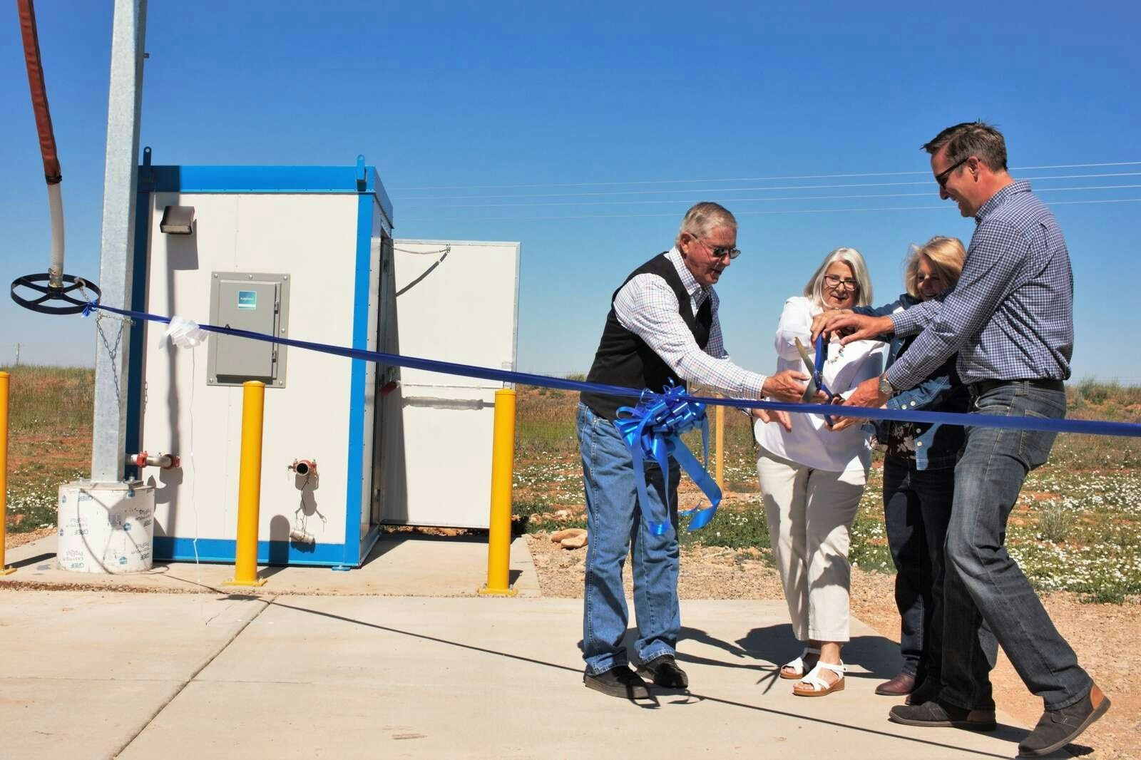 Four people are cutting a blue ribbon at an outdoor event, likely a ceremony or opening of a facility.