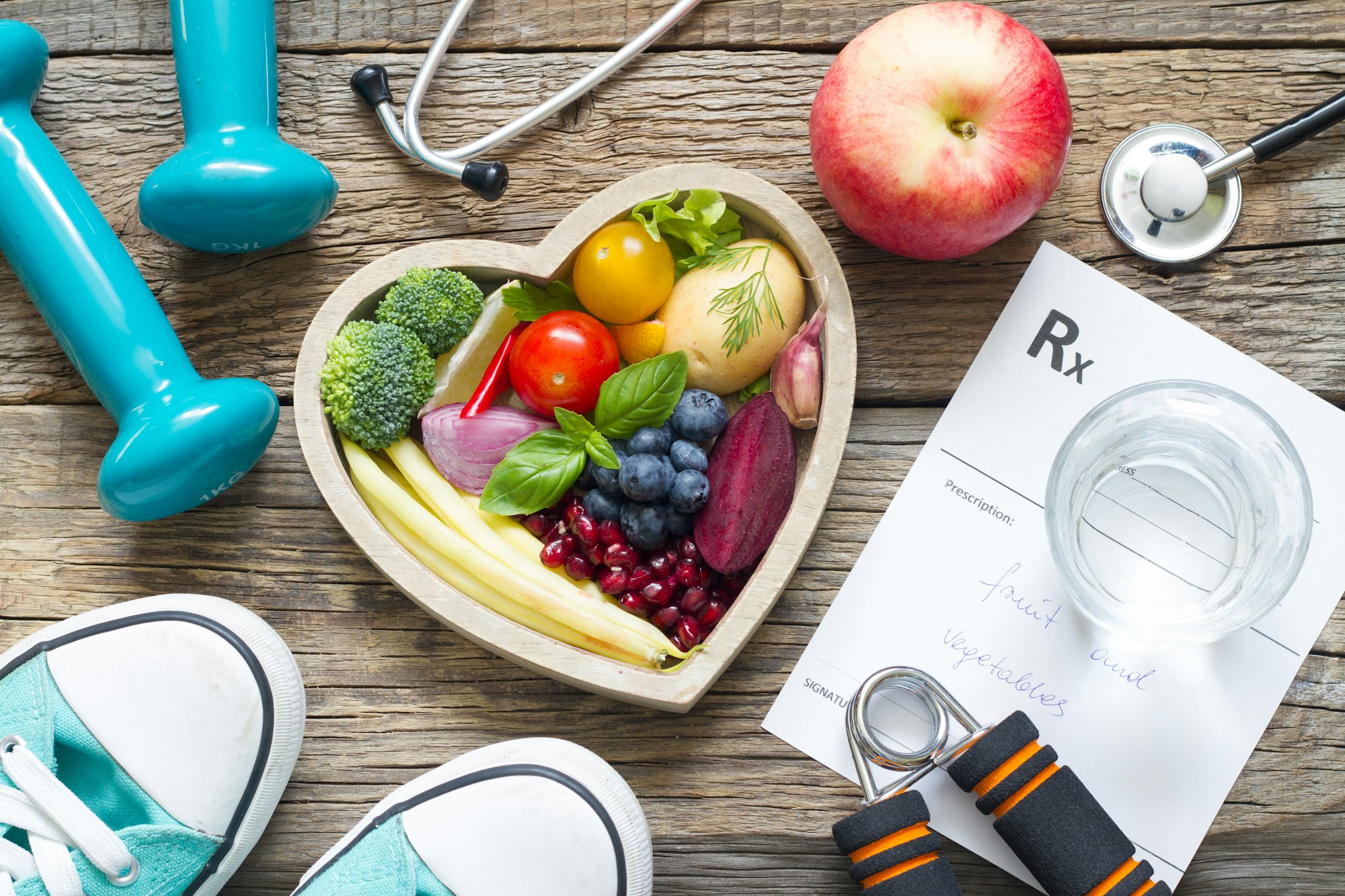 A heart-shaped bowl with fresh produce, dumbbells, sneakers, stethoscope, prescription for fruits/vegetables, glass of water on wooden surface.