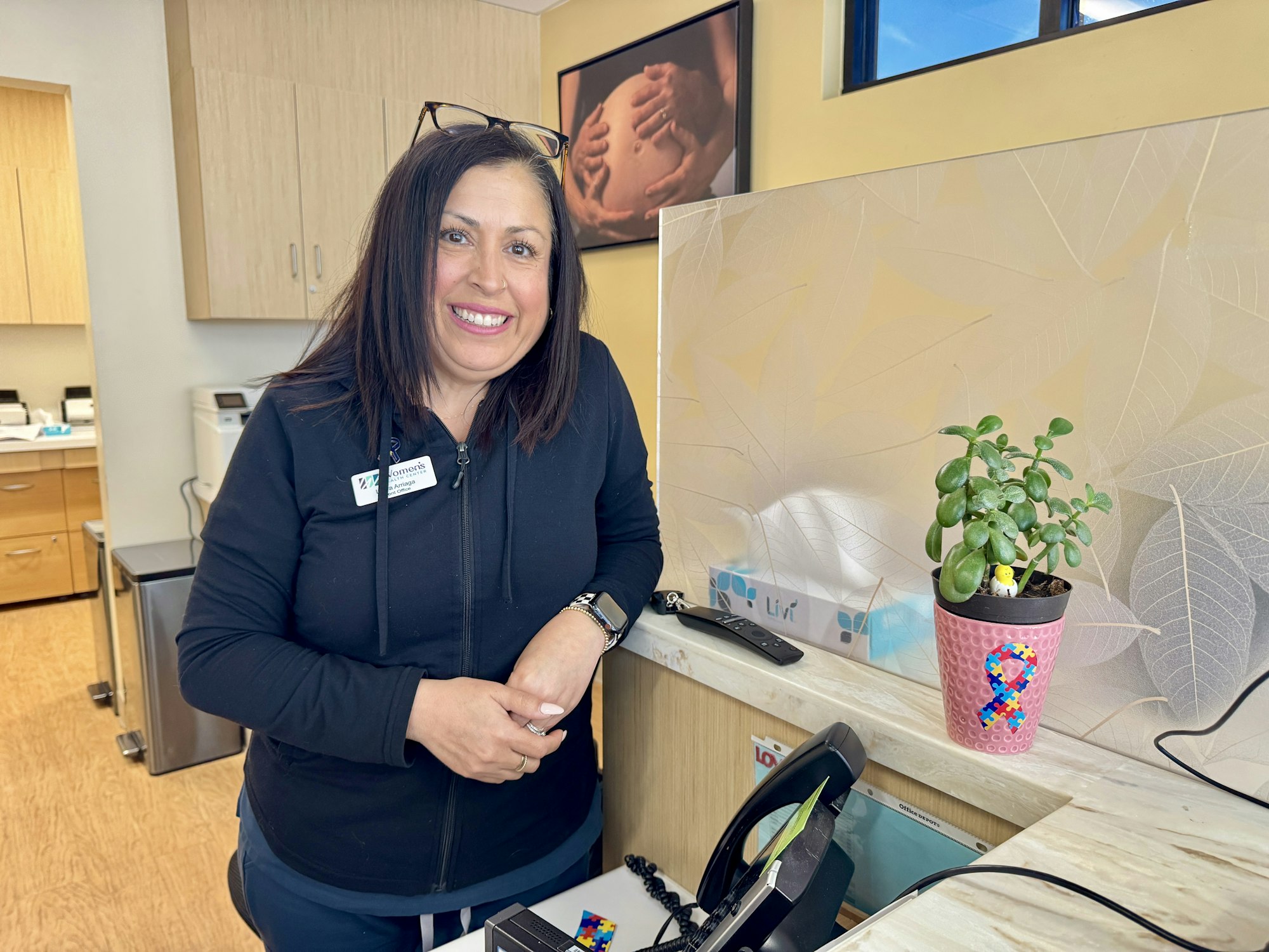 A person smiling at a reception desk with a plant in a pot, featuring a puzzle piece pattern, and a phone nearby.