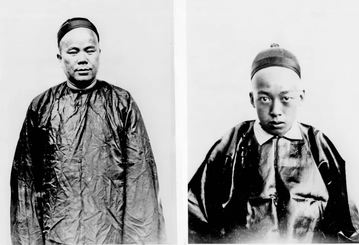 The image shows two individuals, both wearing traditional dark garments and caps, posing against a plain background.
