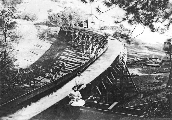 An old black-and-white photo of two women by a wooden flume or slide, set in a rural landscape, with trees and structures nearby.