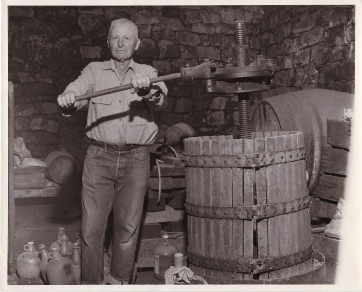 A man operating a traditional wine press in a rustic cellar, surrounded by barrels and bottles.