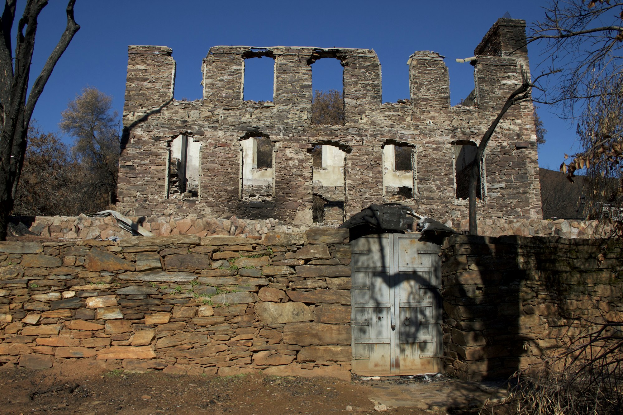 A weathered stone ruin with missing windows, a stone wall, and a blue metal door, set against a clear blue sky.