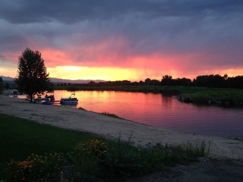 Sunset over a river with boats docked along the shore, silhouetted trees, and vibrant orange and pink skies.