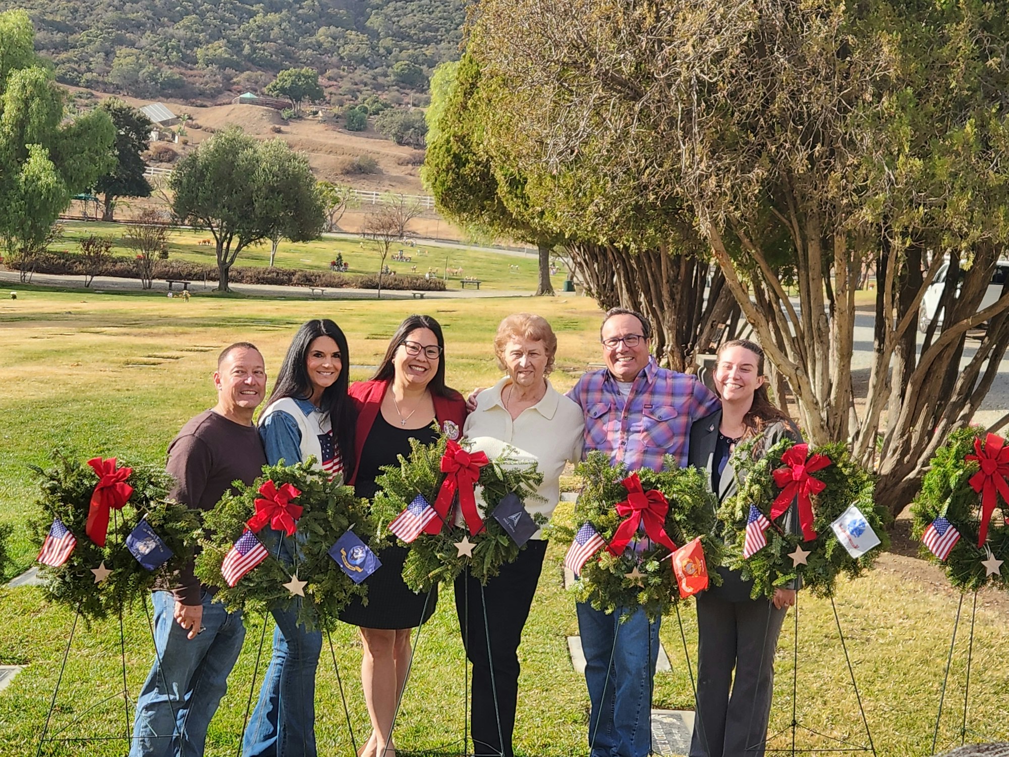 A group of six people smiling, standing outdoors with wreaths adorned with red bows and small flags.