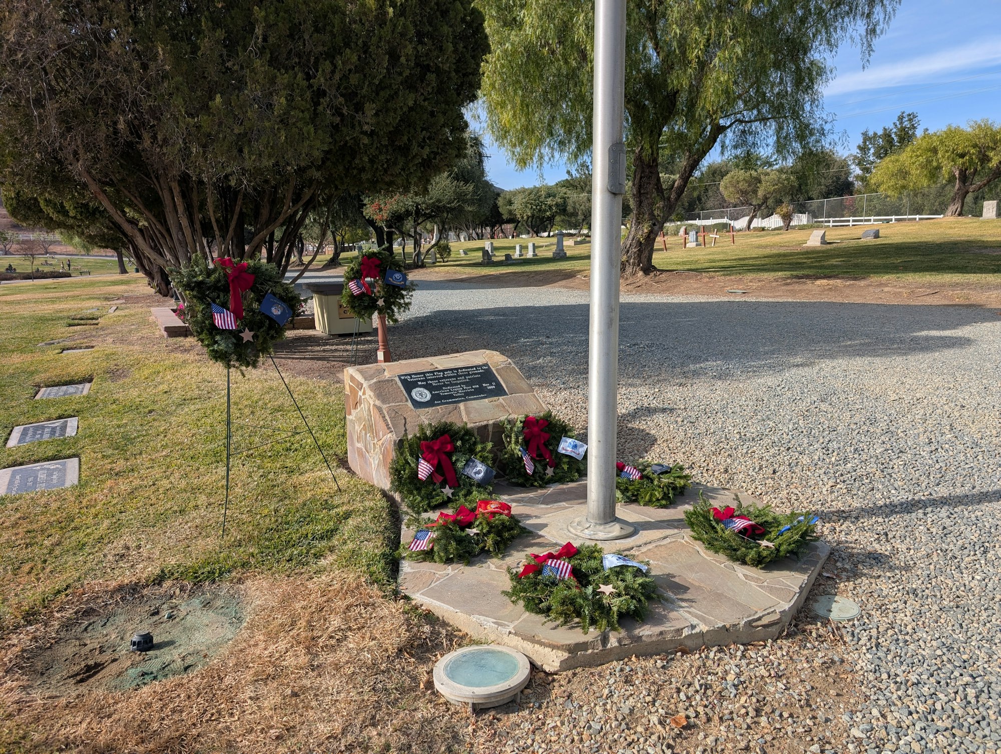 Wreaths with flags and bows are placed around a flagpole and memorial plaque in a cemetery.