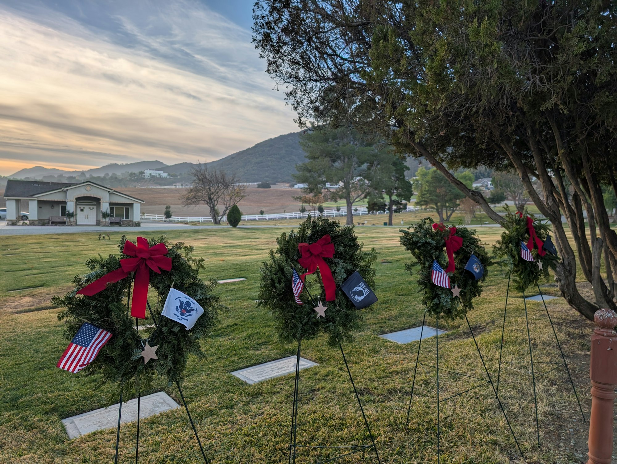 Wreaths with flags and bows on stands in a cemetery with a mountain view and house in the background.