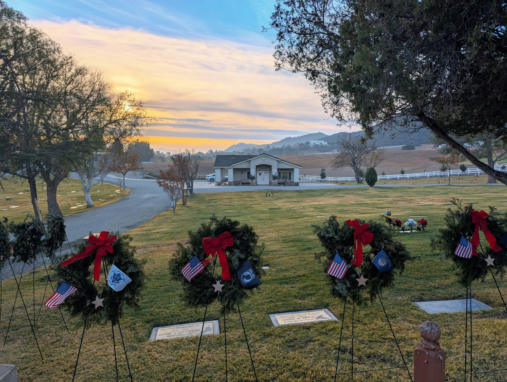 Wreaths with flags and bows in a cemetery at sunset, with a building and hills in the background.