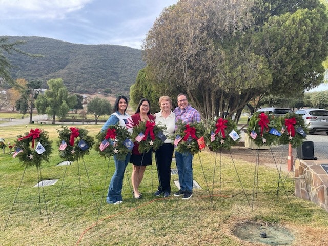 Four people stand together outdoors, holding wreaths with red bows, surrounded by trees and mountains in the background.