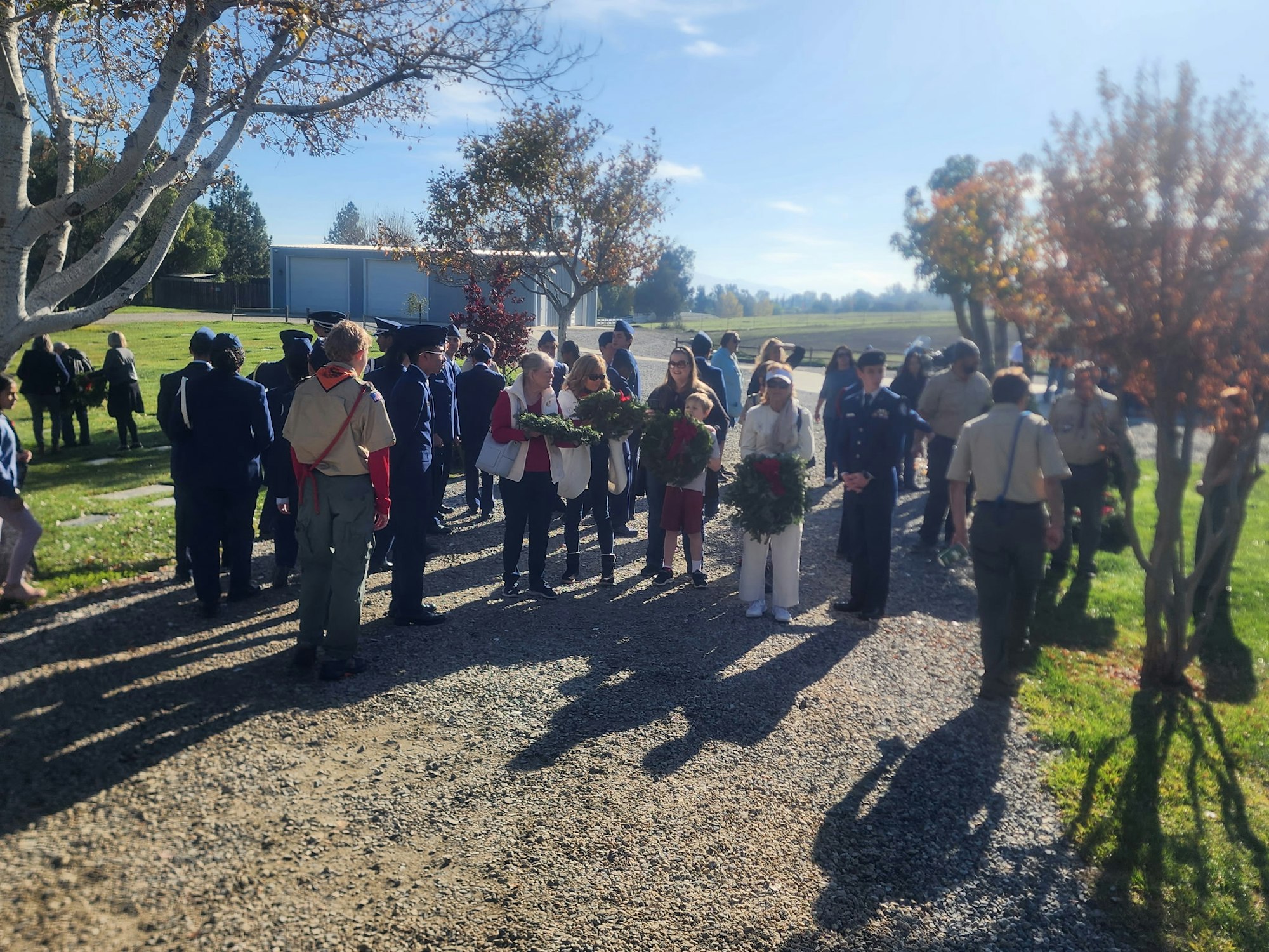 A group of people gathered outdoors, some in uniforms, holding wreaths under a clear sky, likely for a memorial or ceremony.