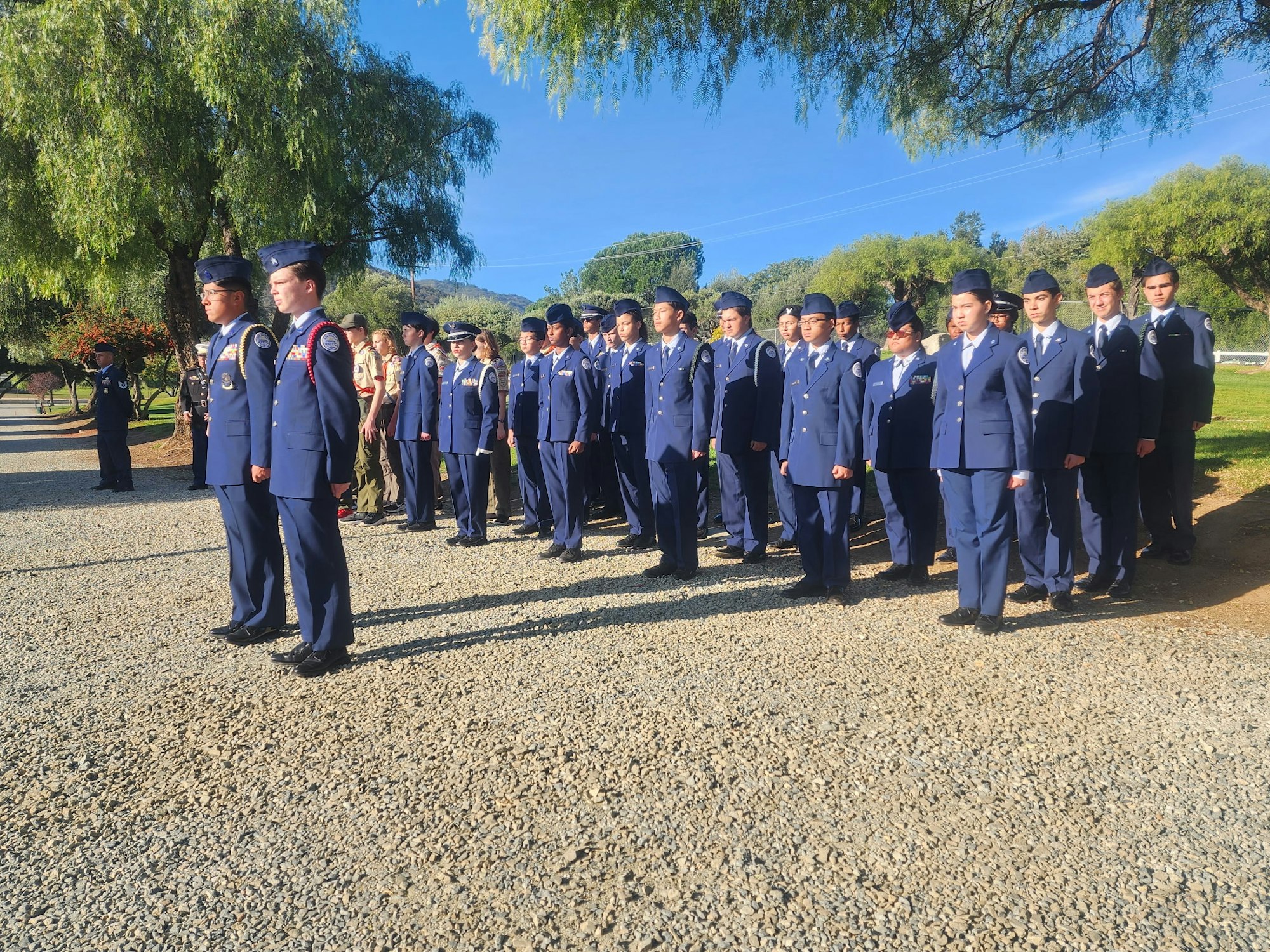 A group of uniformed cadets stands in formation outdoors, surrounded by trees and a clear blue sky.