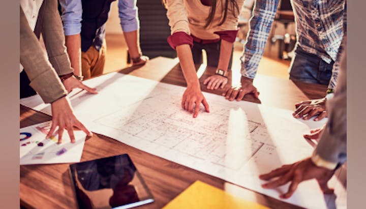 A group of people gathered around a table, discussing and pointing at architectural plans.