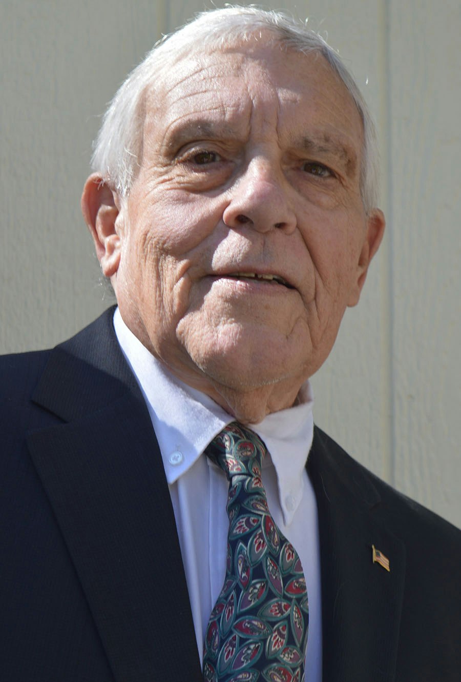The image shows an older man in a suit and tie, with a subtle smile and an American flag pin on his lapel.